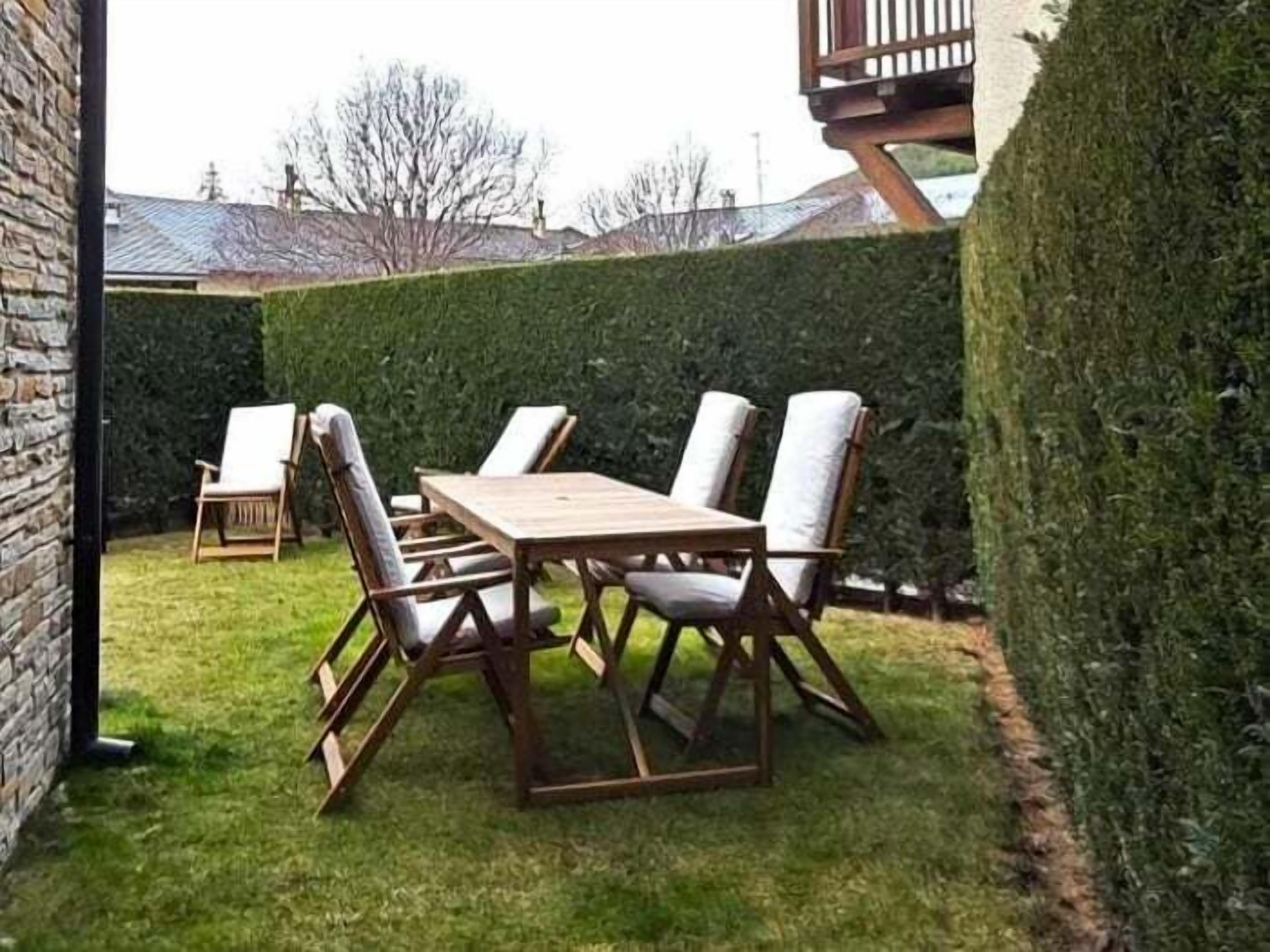 Une table et des chaises en bois, installées dans un jardin agrémenté d’une haie et d’un mur en briques.