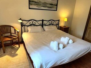 Un lit bien fait, avec du linge de lit et des serviettes blanches, une chaise en bois, une table de chevet avec une lampe et un tableau accroché au mur.