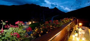 Eine Terrasse mit Holzbalkon, ein Tisch mit Weingläsern und einer Flasche, sowie ein Blick auf die Berge im Hintergrund.