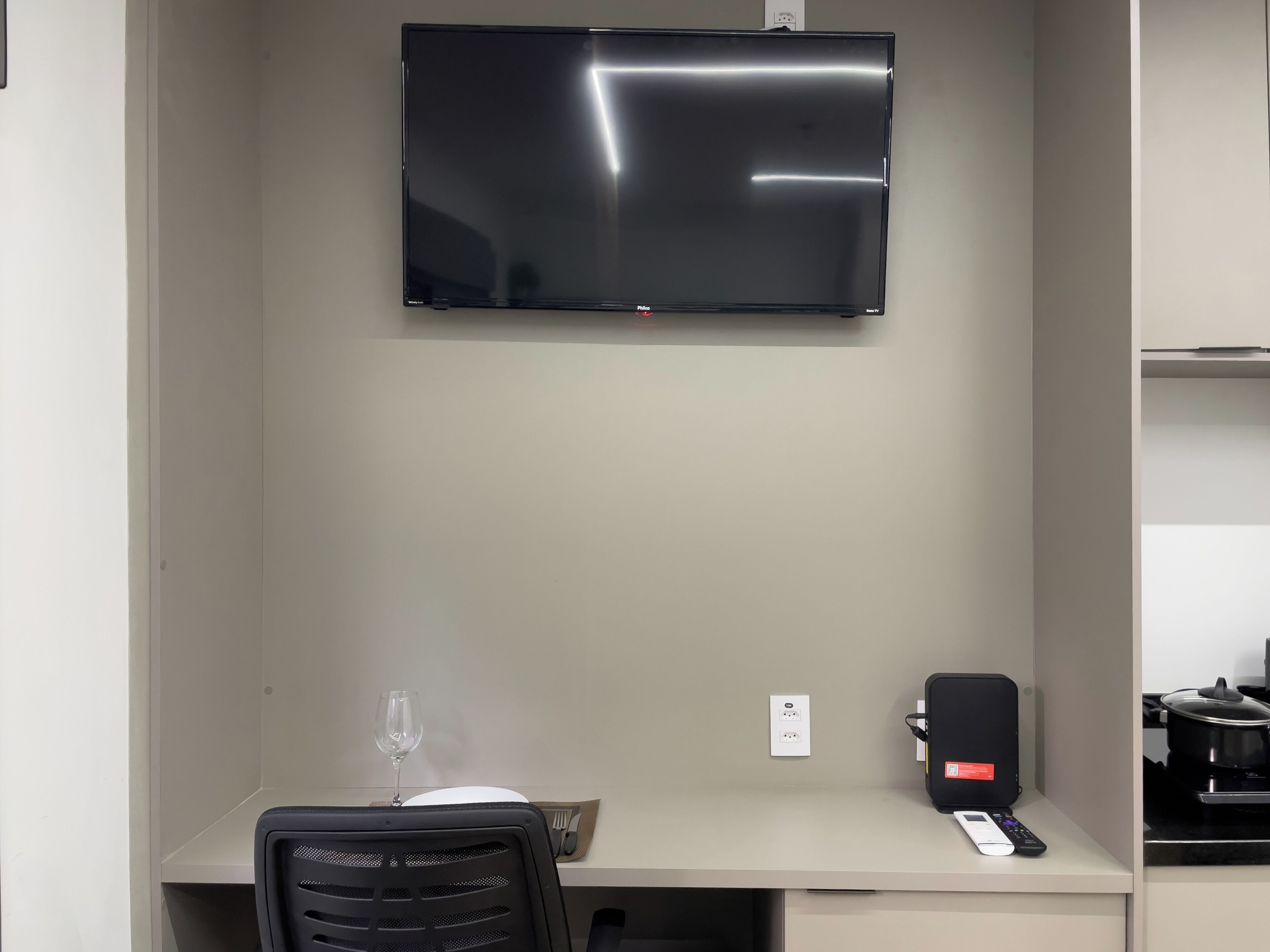 Ein Hotelzimmer mit Flachbildfernseher an der Wand, einem Schreibtisch mit Stuhl, einem Weinglas, einem Lautsprecher und einer Fernbedienung.