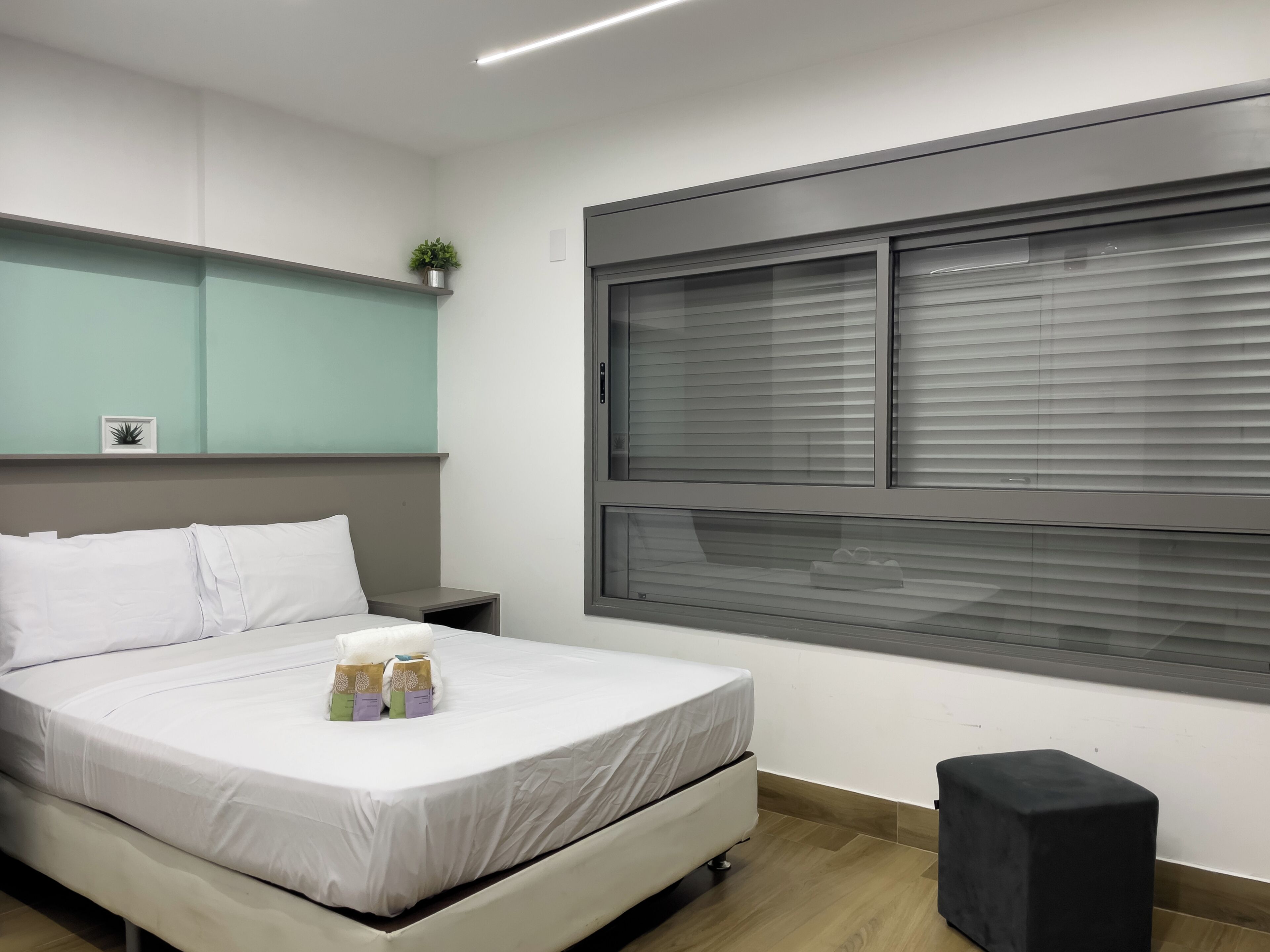 A modern bedroom with a bed, bedside table, window with blinds, and a small potted plant.