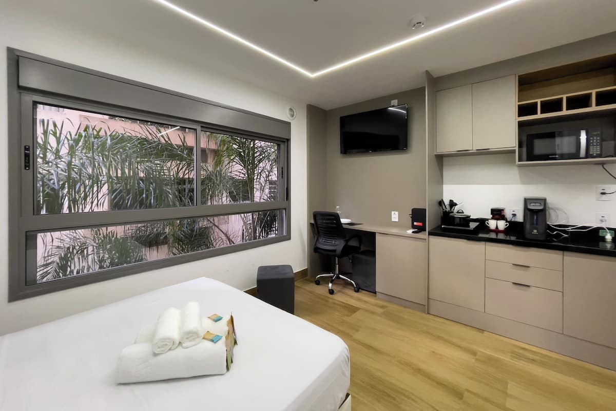 A modern room with a bed, a desk, a TV, and a kitchen area.