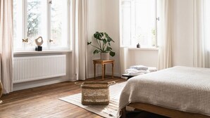 Une chambre avec un plancher en bois, un lit recouvert d’une couette à carreaux, une plante en pot, une petite table en bois et un panier.