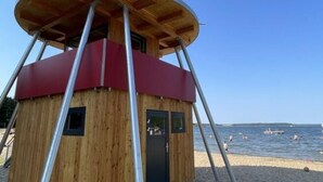 Une cabane de plage en bois, avec un toit rouge et des supports métalliques, située sur une plage de sable, avec la mer en arrière-plan.