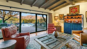 A spacious living area with leather sofas, a wooden coffee table, and a view of a pool and mountains through large sliding glass doors. - Private Foothills Villa + Panoramic Views + Heated Pool/Spa (Tucson)