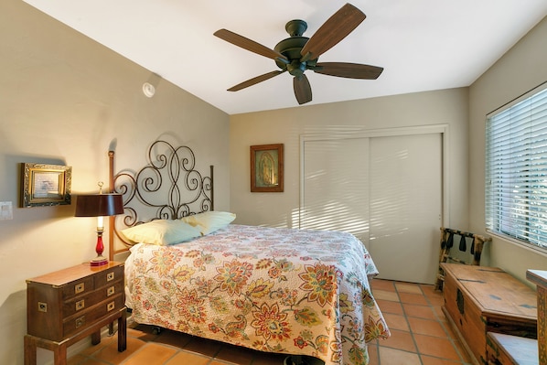 Une chambre à coucher comprenant un lit, un ventilateur de plafond, une table de chevet, une commode, une fenêtre avec des stores et une porte.