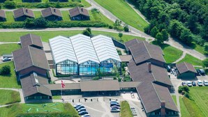Eine Vogelperspektive zeigt einen Gebäudekomplex mit einem zentralen Schwimmbecken, umgeben von mehreren kleineren Gebäuden, einem Parkplatz und grünen Rasenflächen.