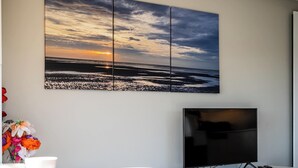 A room with a large wall-mounted TV, a white console table, and a multi-panel artwork depicting a sunset over the ocean. - Apartment in Oostduinkerke Near the Beach (Koksijde)