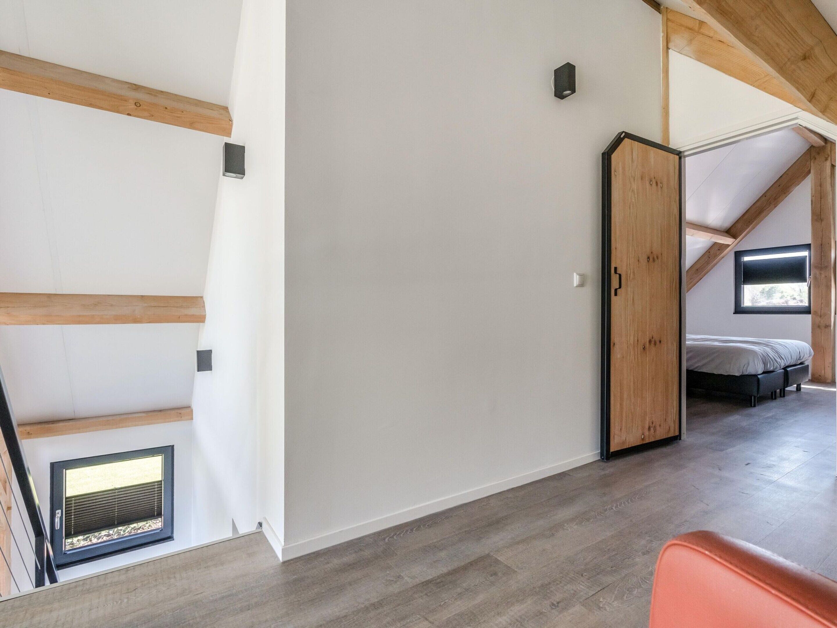 A room with a sloped ceiling, wooden beams, a bed, a small window, and a door.