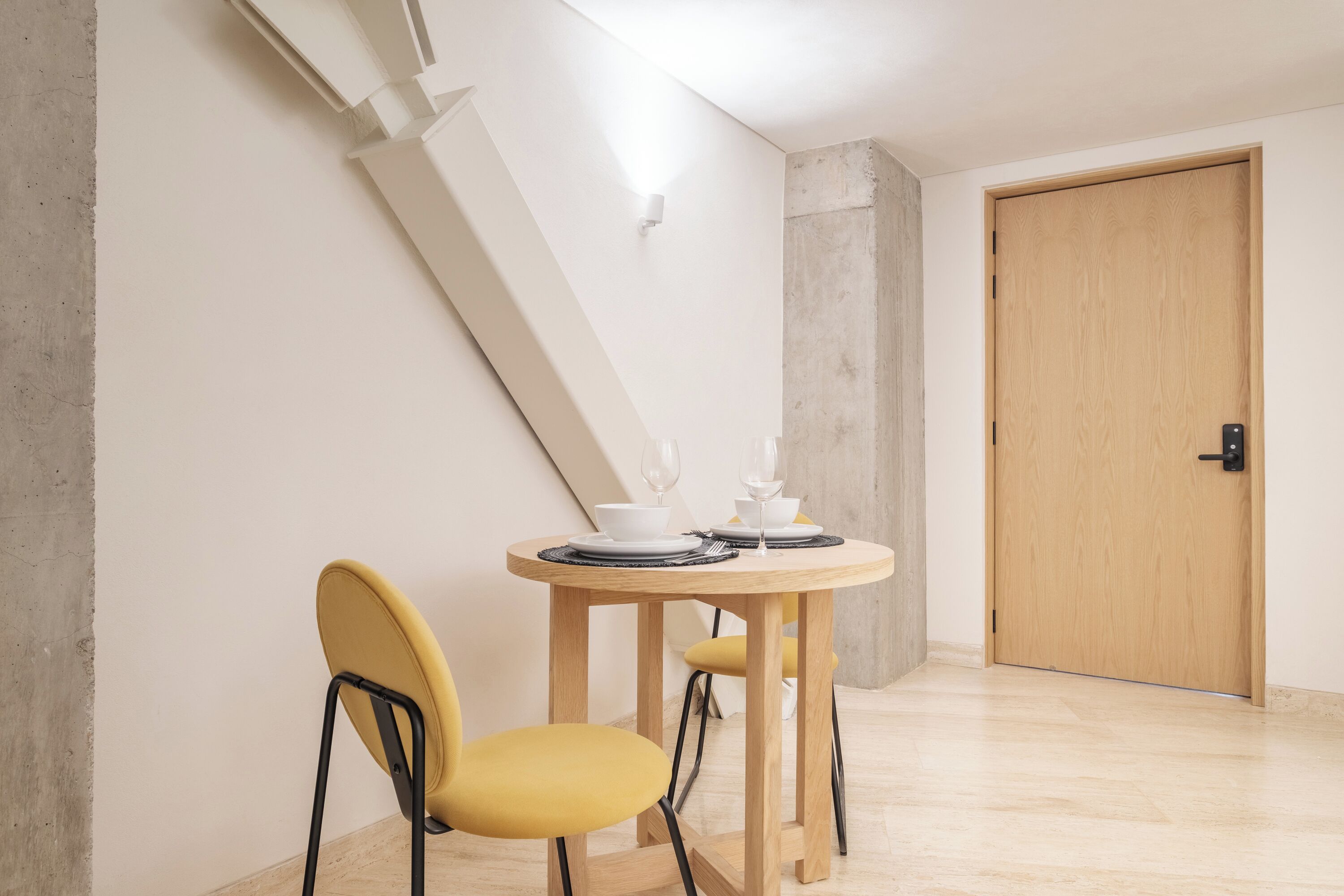 Een eetruimte met een ronde houten tafel, twee gele stoelen en een witte muur met een lamp.