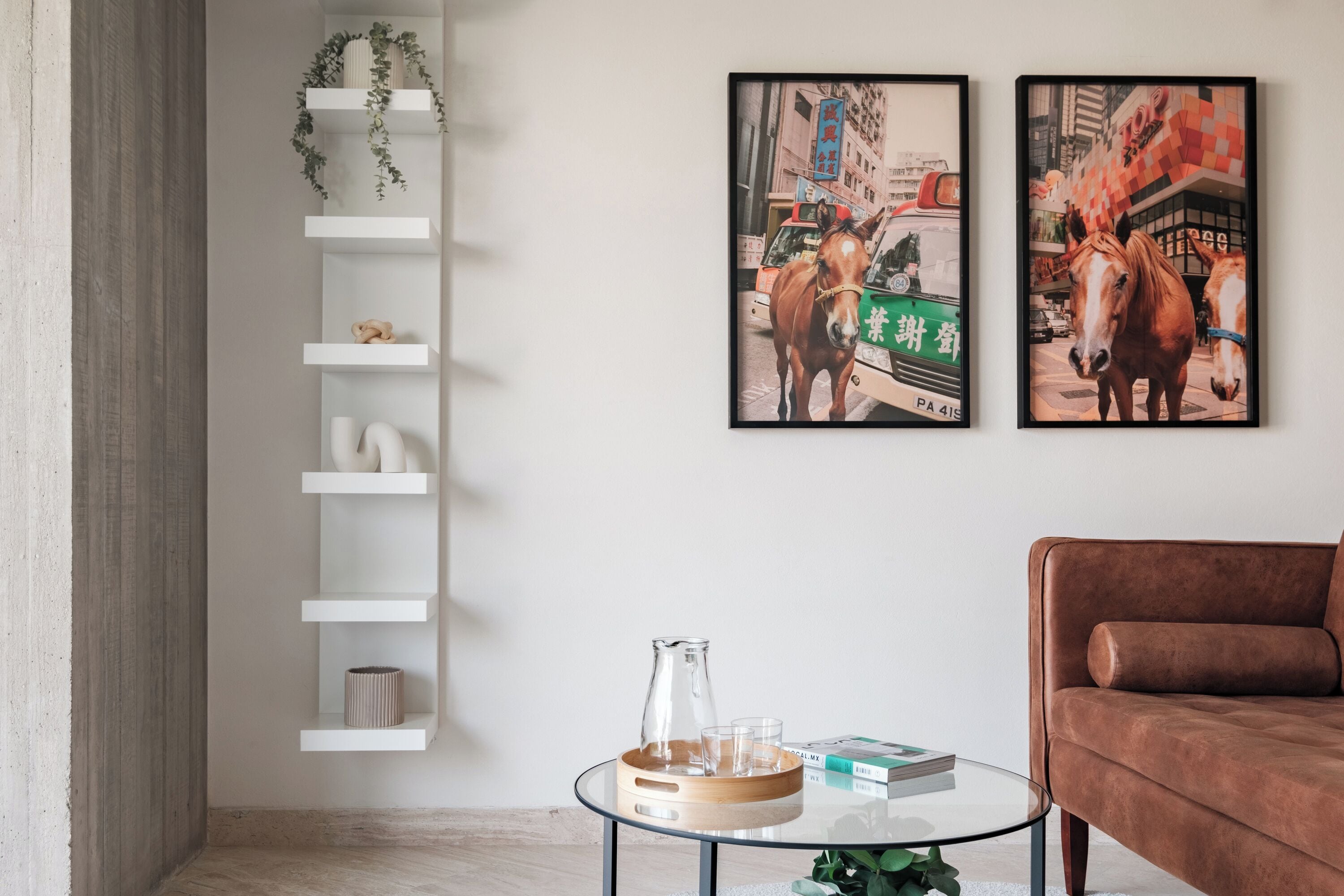 Un salon moderne avec une table basse en verre, un canapé marron et deux tableaux représentant des chevaux, encadrés et accrochés au mur.