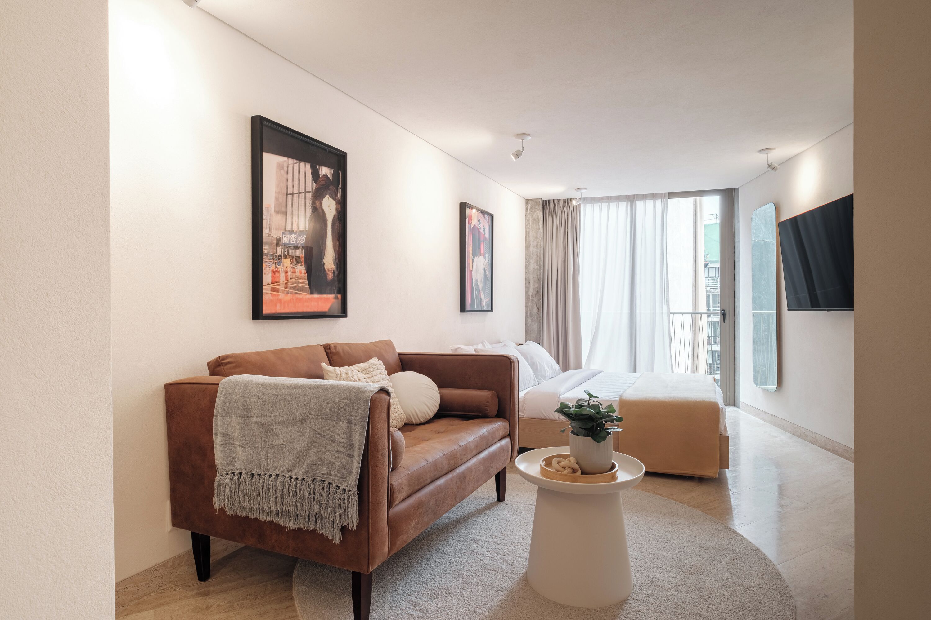Una moderna camera d'albergo con un divano in pelle marrone, un tavolino rotondo e un letto con copripiumino color beige. Si notano due opere d'arte incorniciate al muro e un'ampia finestra con tende.
