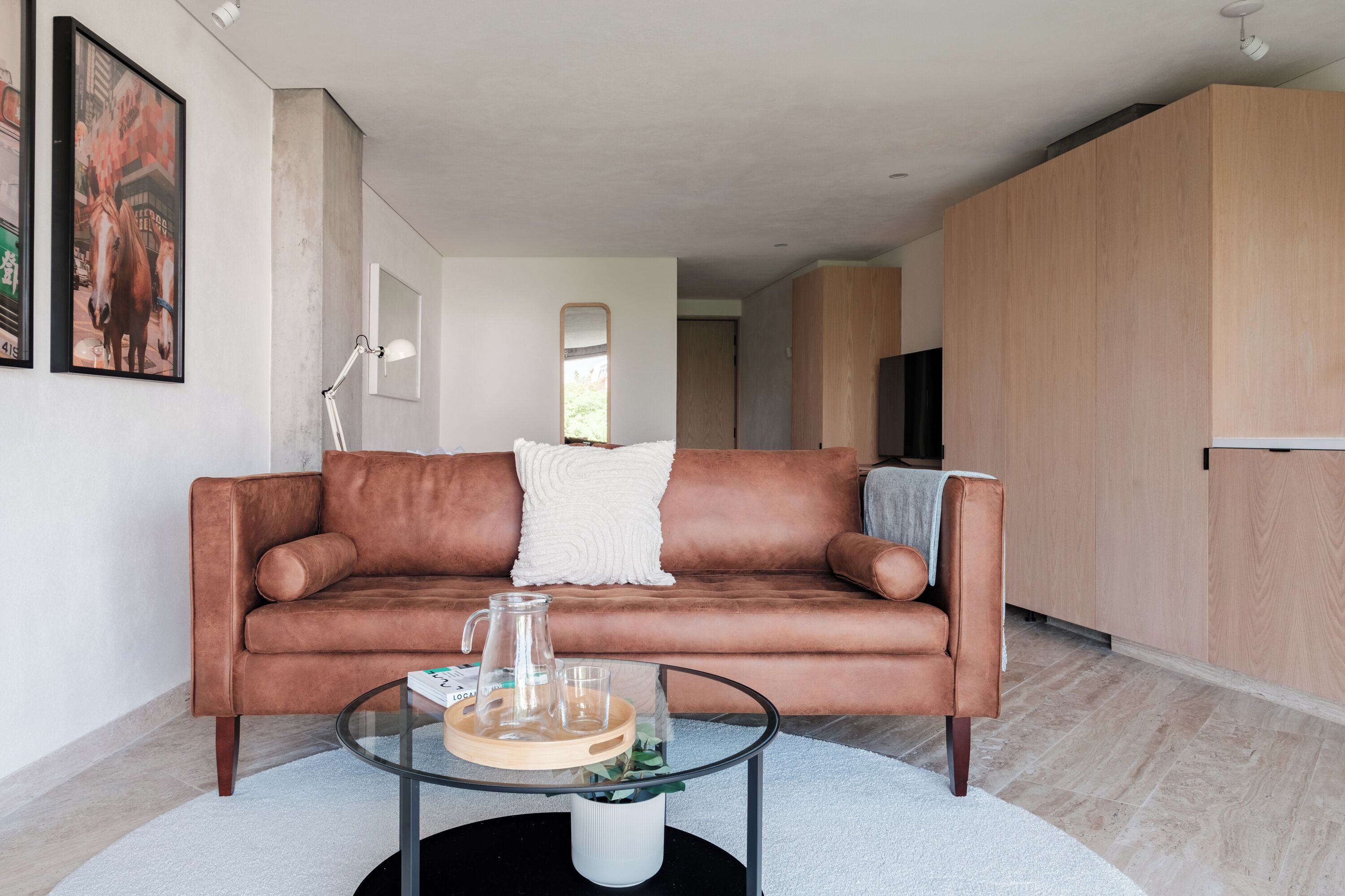 En moderne stue med en lædersofa i brunt, et glasbord og en hvid pude. Der er et billede i ramme på væggen og en lampe ved siden af.
