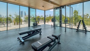 Un gimnasio con equipo de levantamiento de pesas, bancos y vistas a la ciudad a través de amplios ventanales.