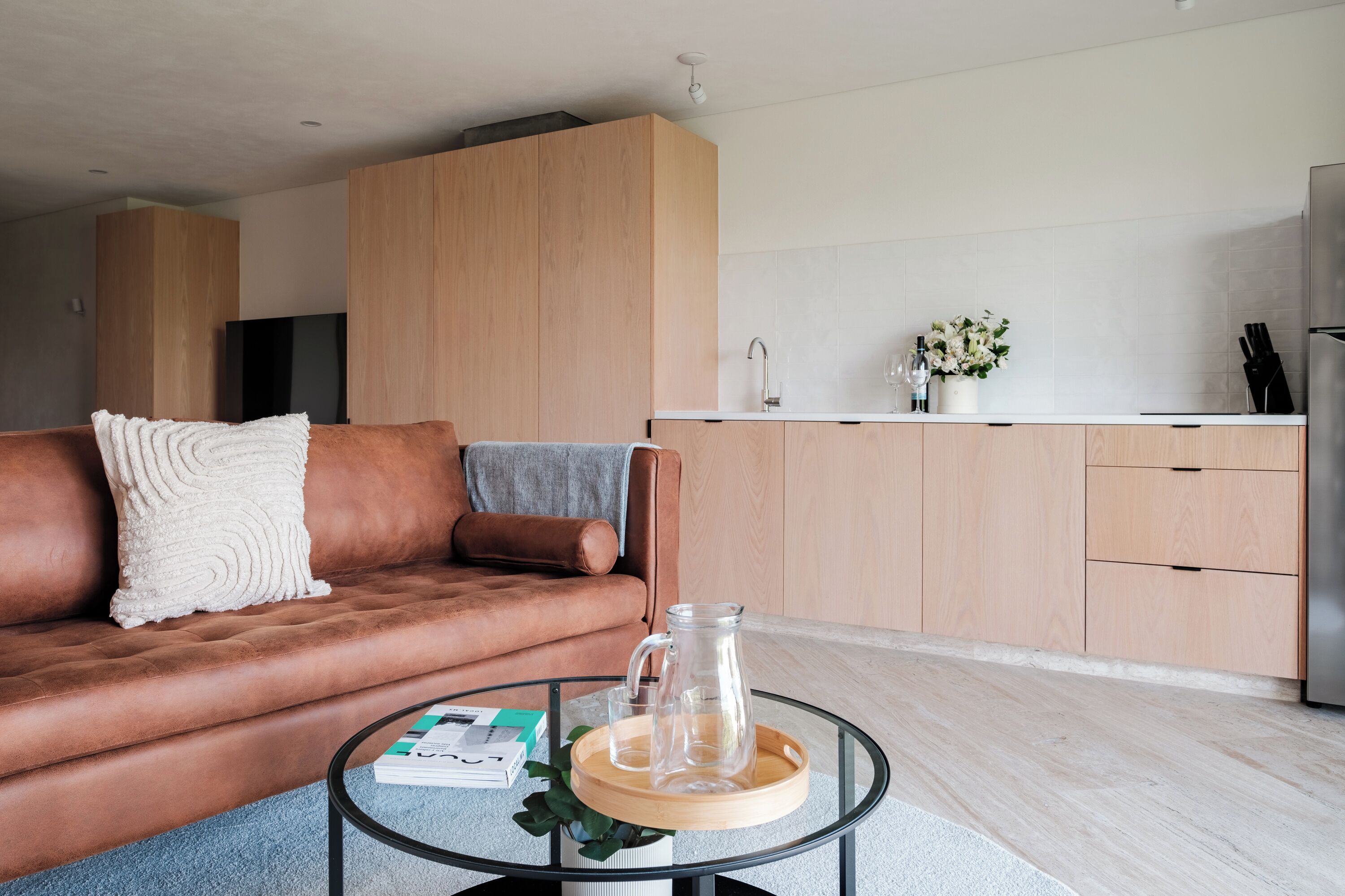 Una sala moderna con un sofá de cuero marrón, una mesa de centro de vidrio y una cocina al fondo.
