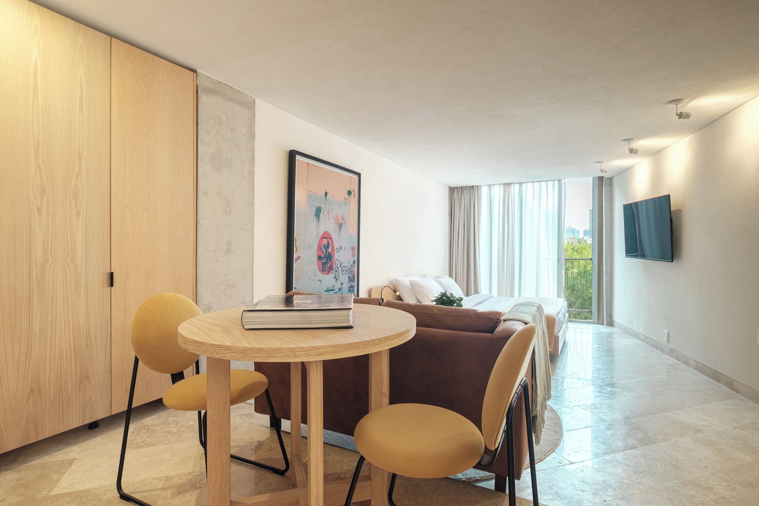 A modern living room with a round wooden table, yellow chairs, a brown sofa, and a TV mounted on the wall.