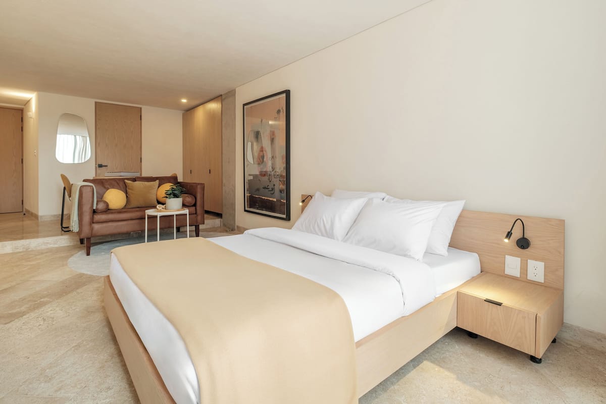 A modern hotel room with a large bed, a small bedside table, a nightstand, a sofa, and a mirror.