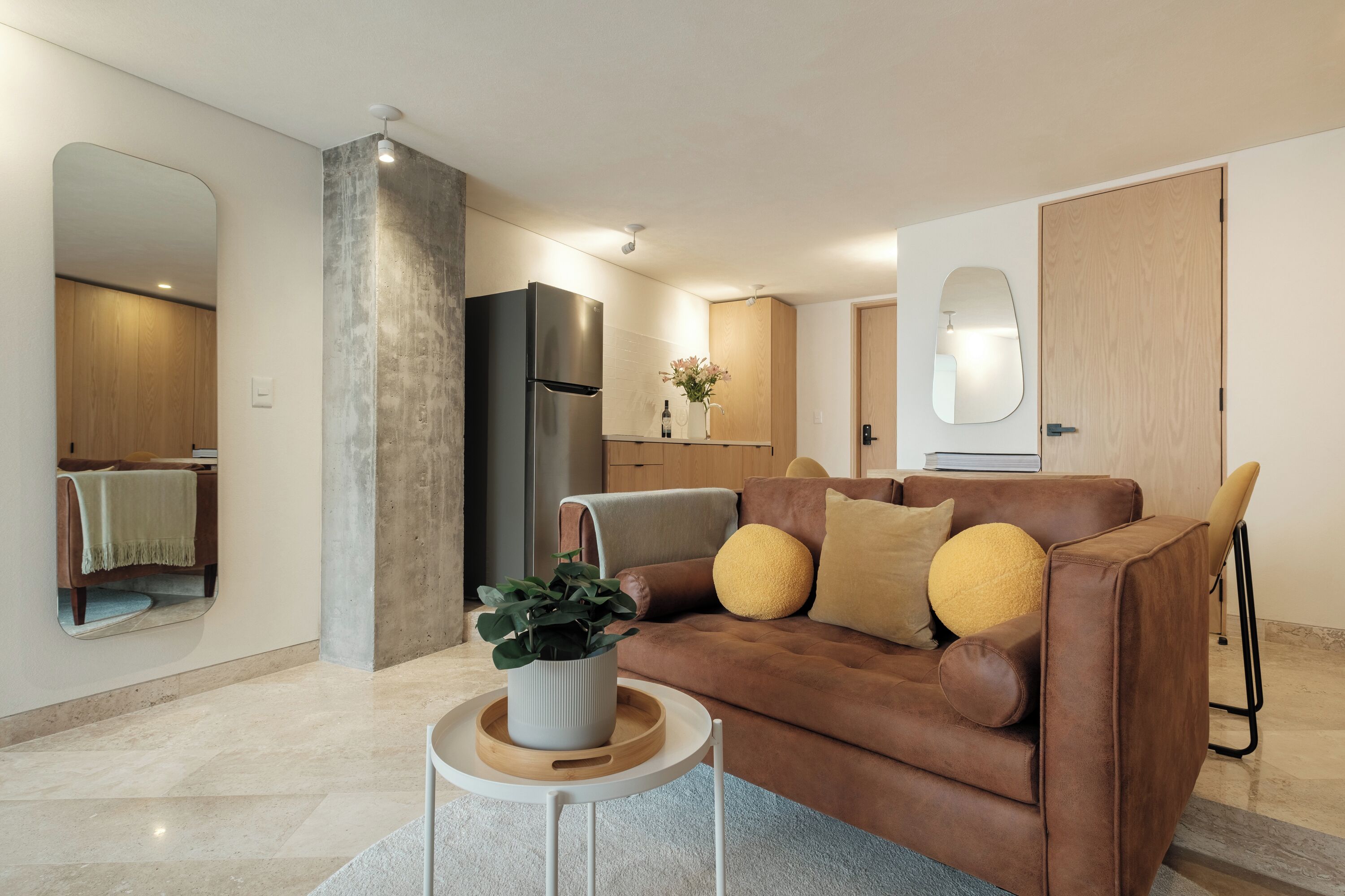 A modern living room with a brown sofa, a small side table with a plant, and a kitchen area in the background.