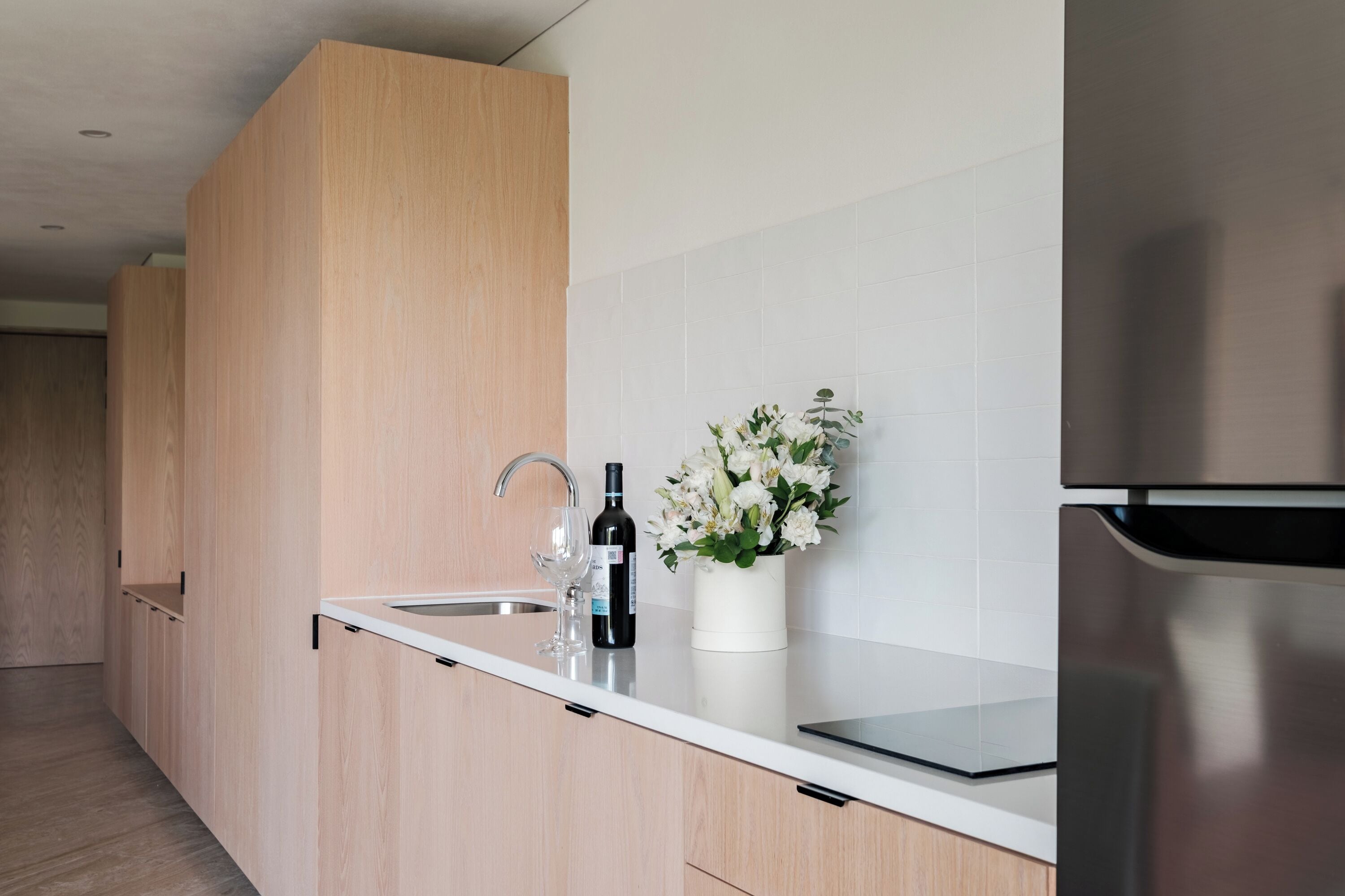 A modern kitchen with wooden cabinets, a stainless steel refrigerator, a wine bottle, and a vase with flowers on the counter.