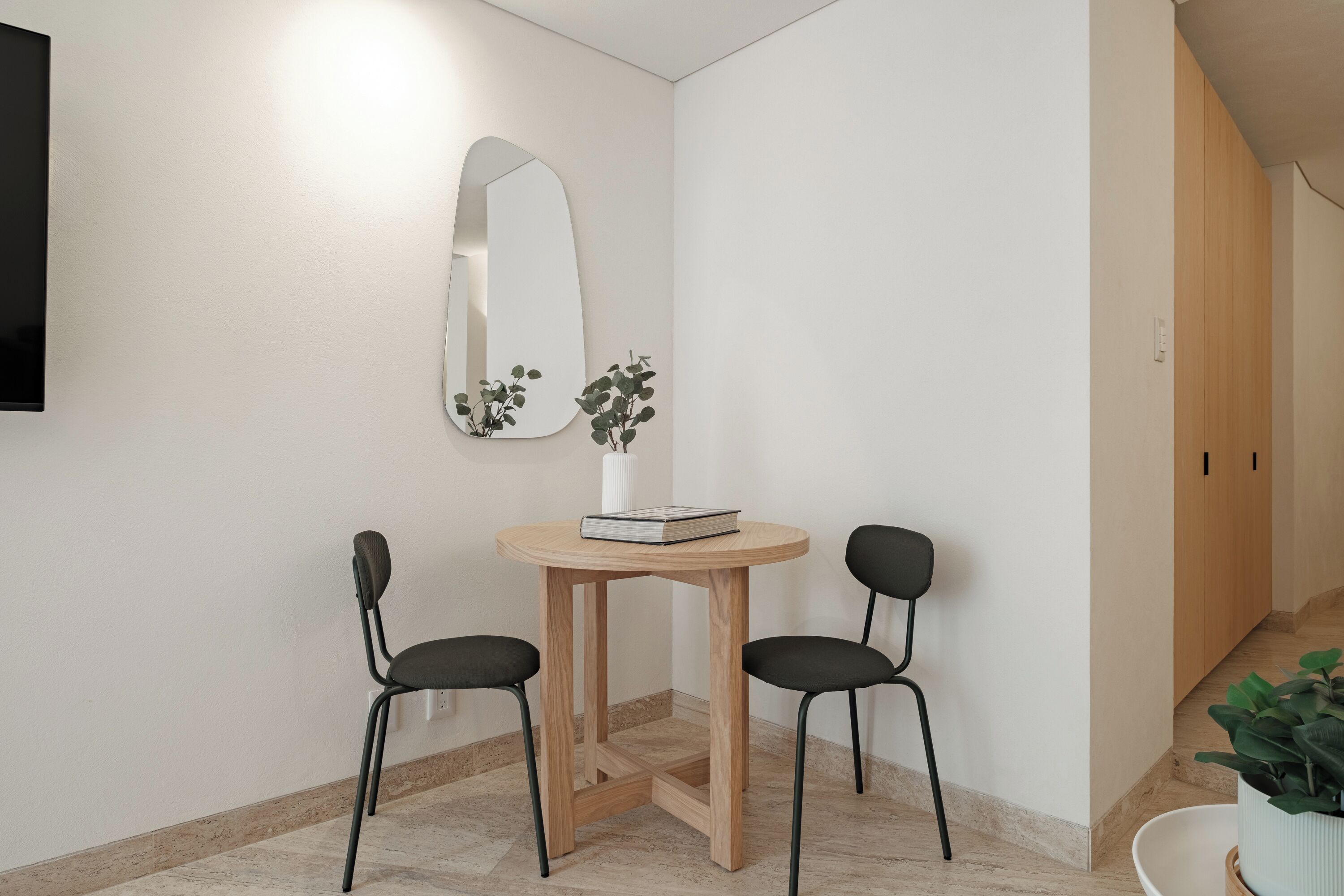 Ein kleiner, moderner Raum mit einem runden Holztisch, zwei schwarzen Stühlen, einer weißen Vase mit Pflanze und einem großen Spiegel an der Wand.
