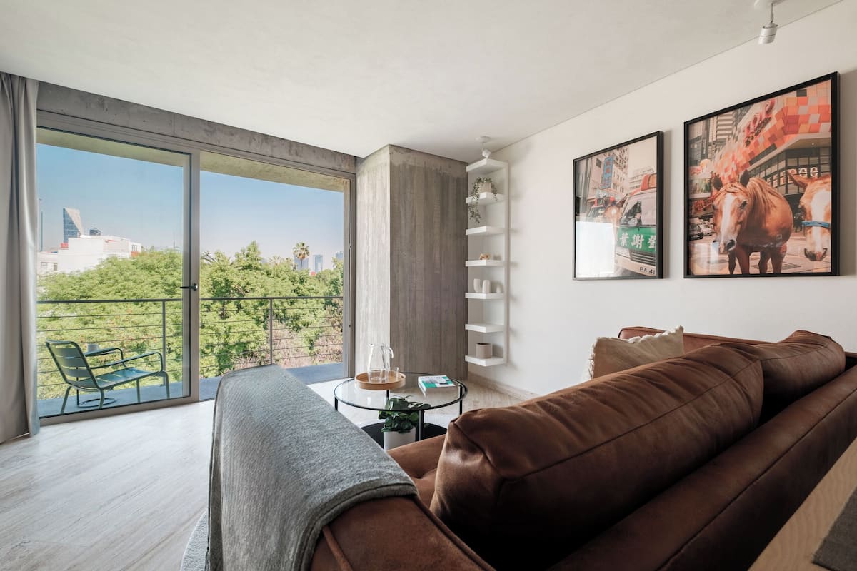 A modern living room with a brown leather sofa, a glass coffee table, and a balcony with a view of a cityscape and greenery.