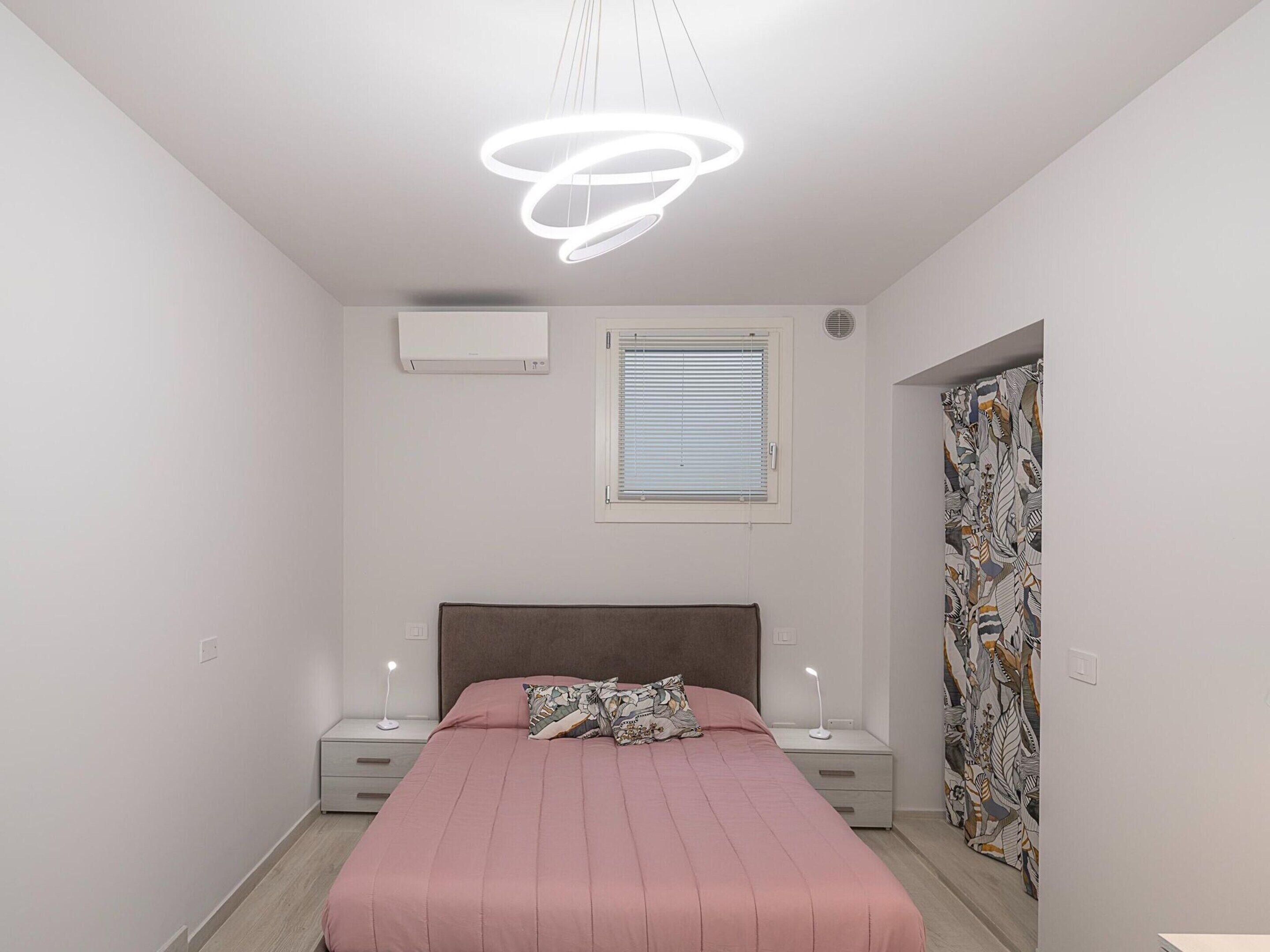 Een slaapkamer met een bed, nachtkastjes, een raam met zonwering, een airconditioning en een gordijn met een bladmotief.