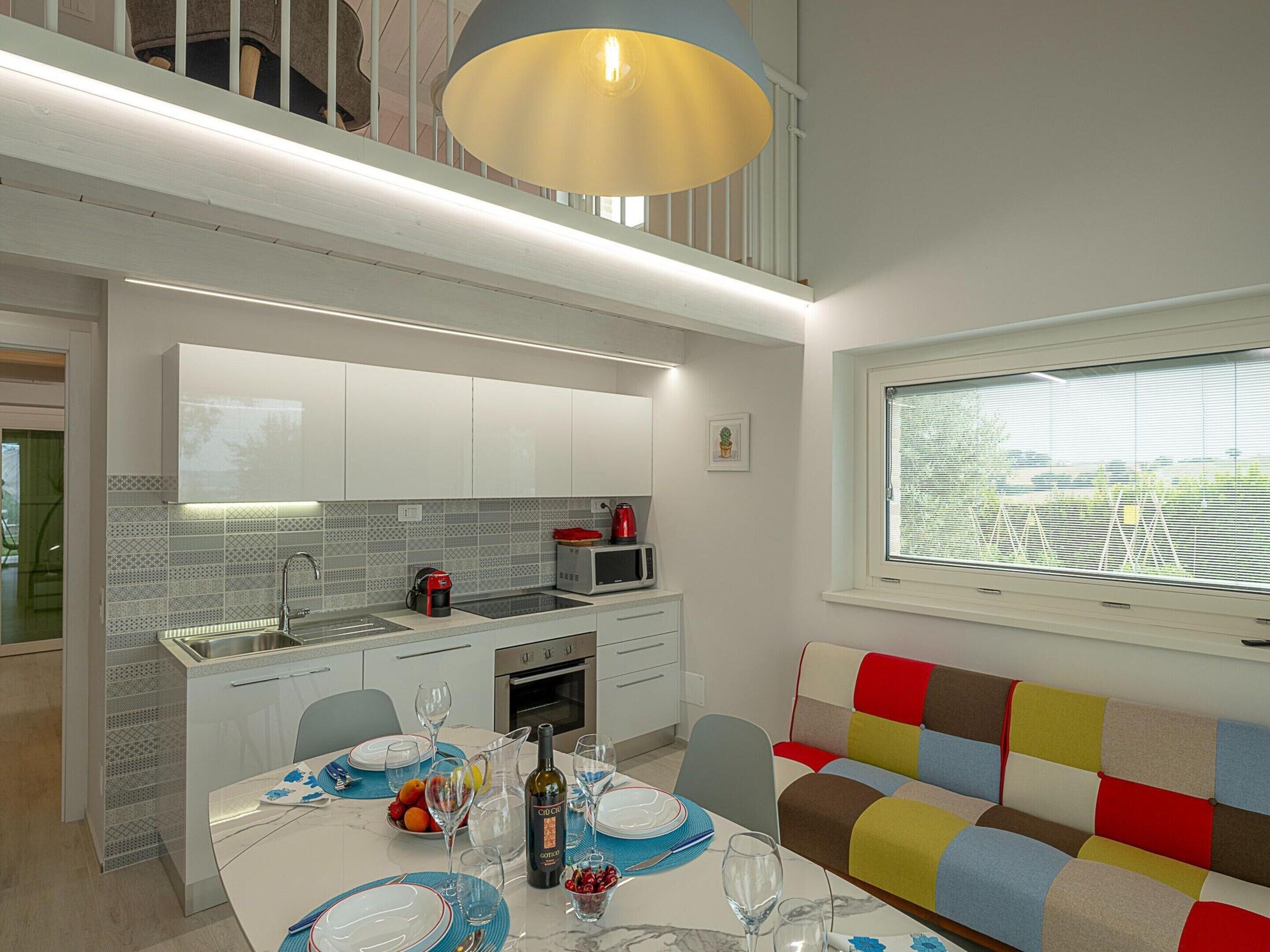 A modern kitchen with white cabinets, a dining area with a table set for two, and a colorful bench.