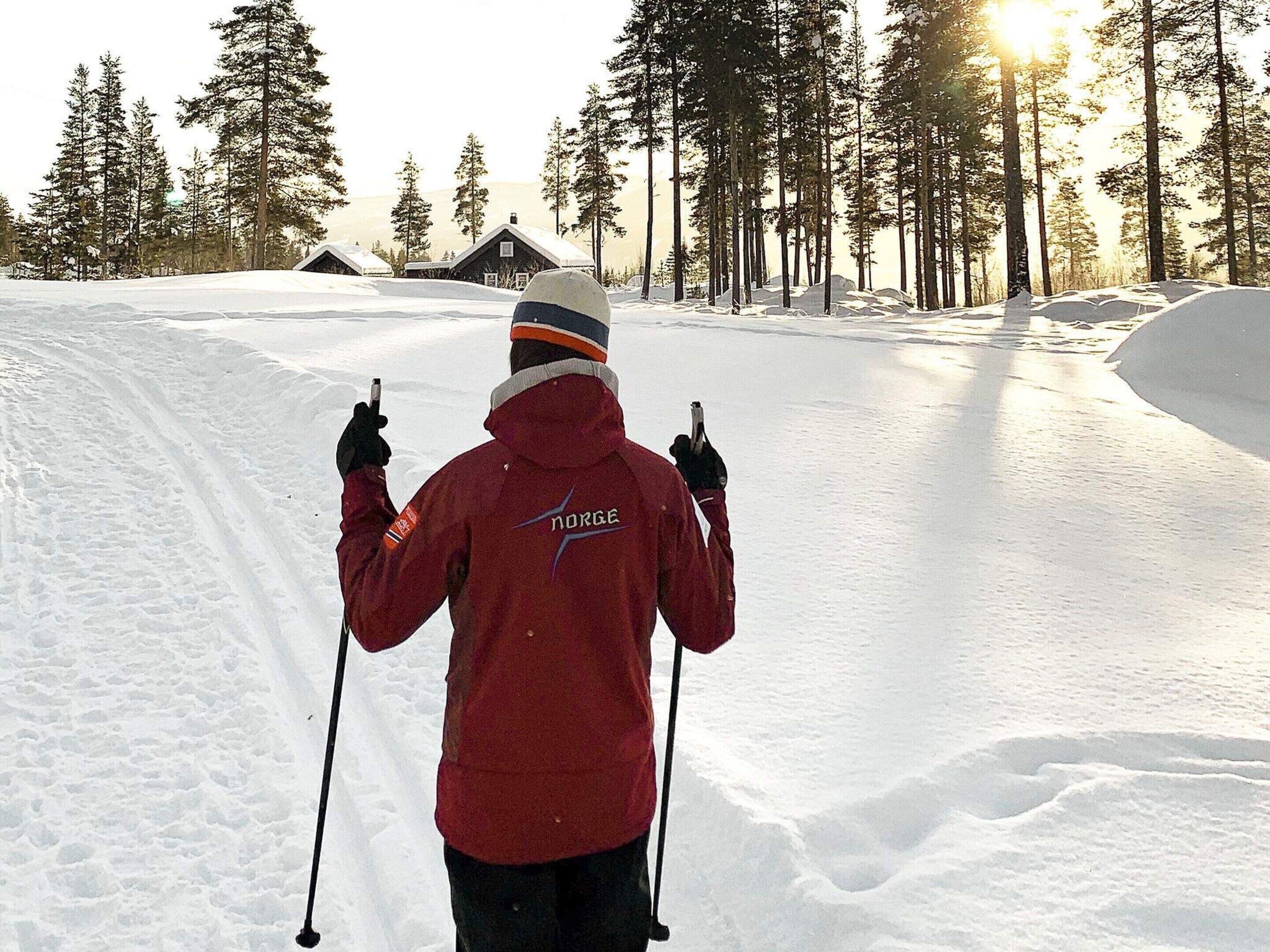 A person in a red jacket with 'Norge' on the back, standing in a snowy area with trees and a cabin in the background.