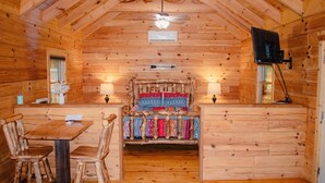 Interior de una cabaña de troncos con una cama de madera, una mesa de comedor con sillas, un ventilador de techo y un televisor montado.