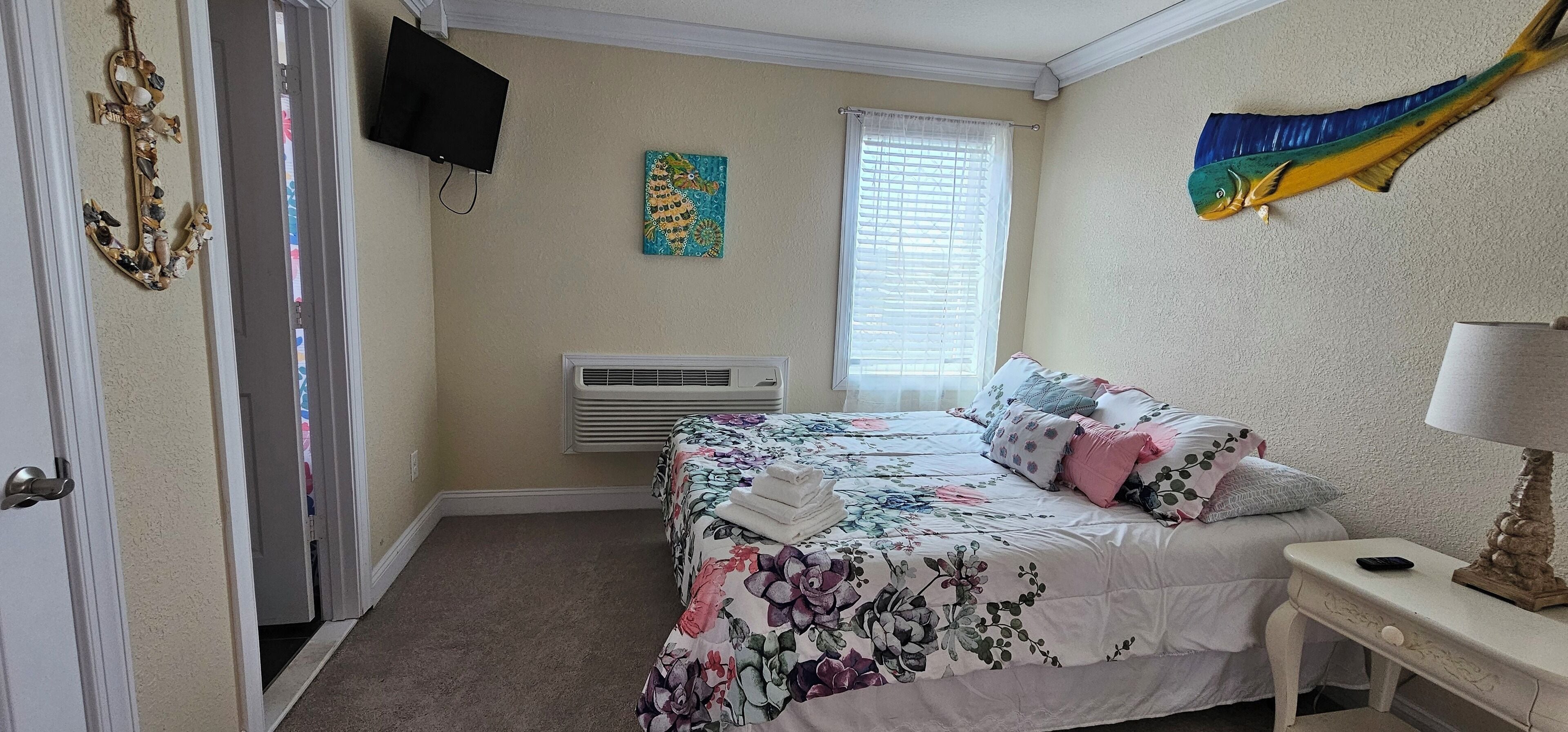 A bedroom with a bed, a bedside table with a lamp, a TV mounted on the wall, a window with blinds, and a decorative fish on the wall.