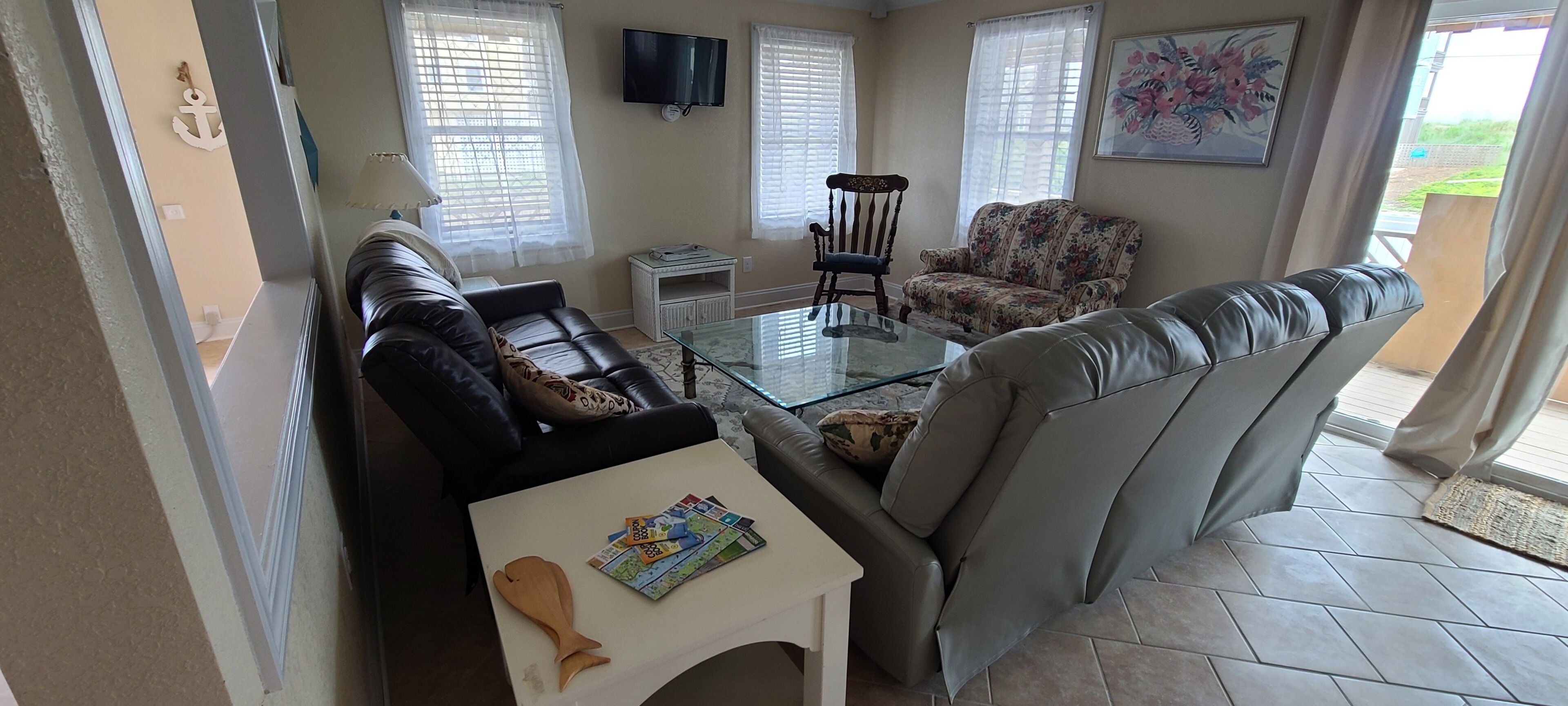 A living room with a sofa, a glass coffee table, a floral armchair, and a television on the wall.