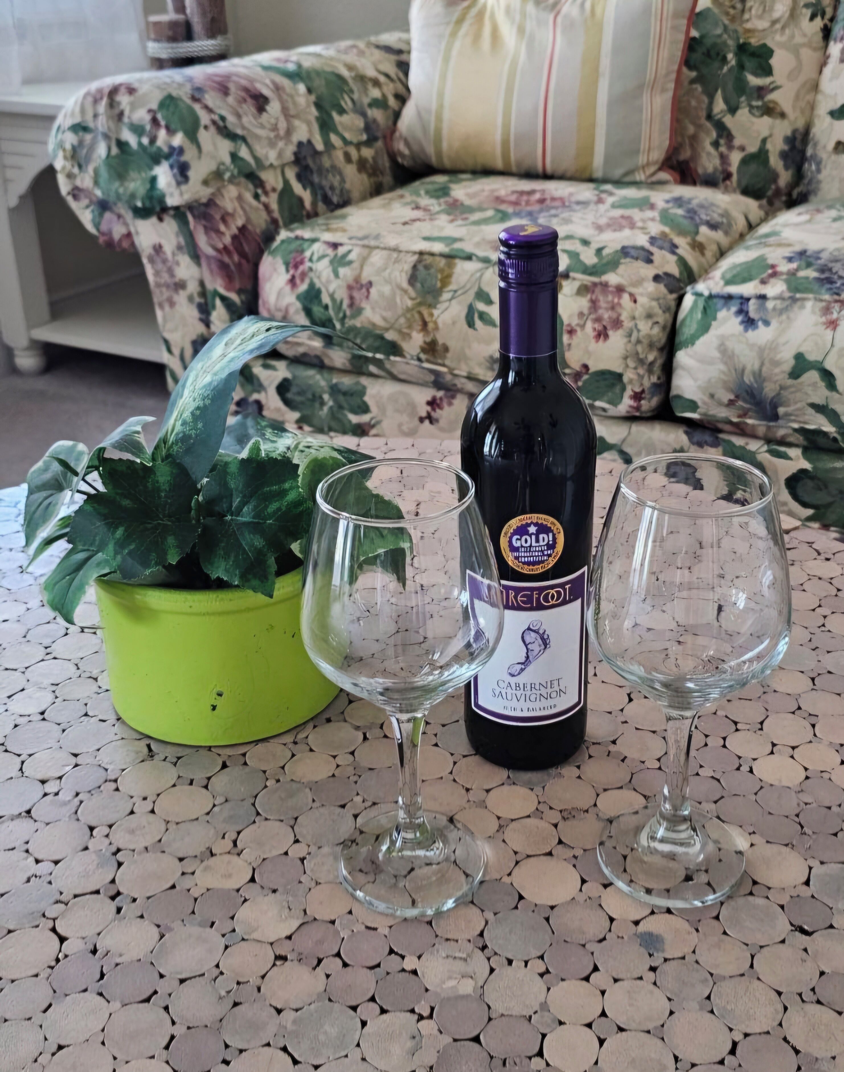 A bottle of wine, two empty wine glasses, and a potted plant on a tiled floor.