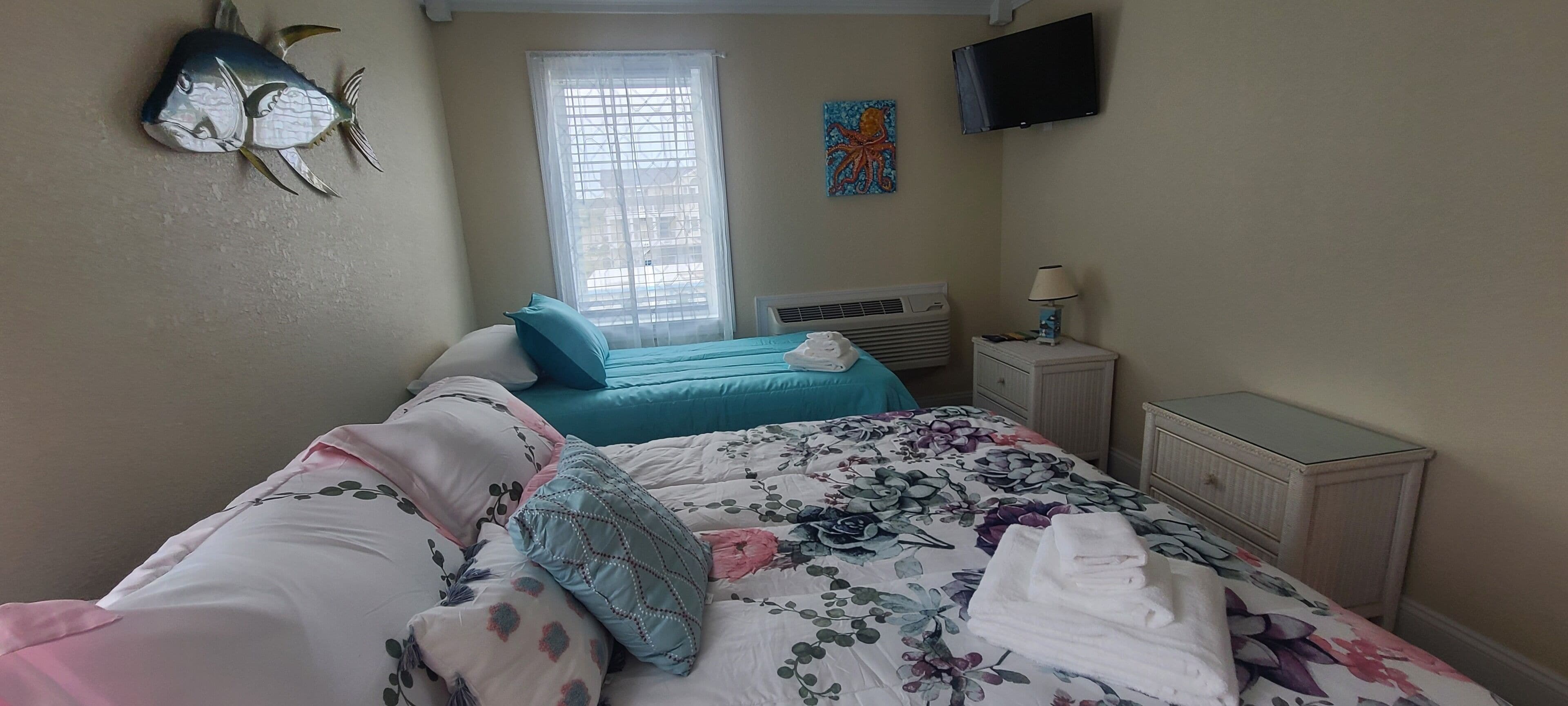 Un dormitorio con dos camas, una ventana con persianas, un televisor de pantalla plana, un cuadro y una mesita de noche con lámpara.