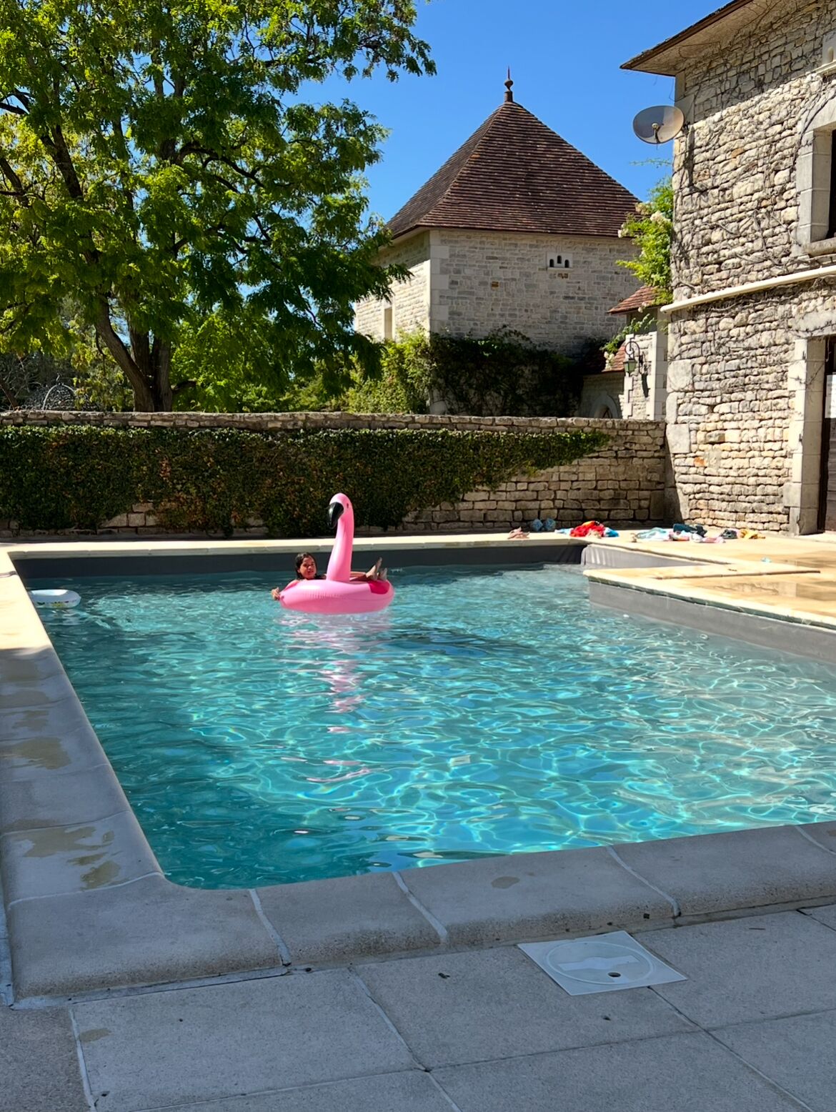 A swimming pool with clear blue water, a pink flamingo float, and stone buildings in the background.