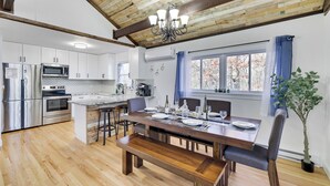 Une cuisine moderne équipée d’appareils électroménagers en acier inoxydable, une table à manger en bois pour quatre personnes et une grande fenêtre agrémentée de rideaux bleus.