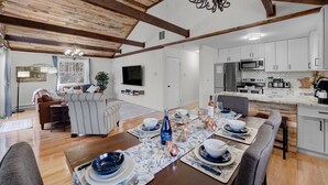 Un vaste salon doté d’un plafond en bois, un coin repas aménagé pour accueillir six personnes et une cuisine moderne avec des meubles blancs et des appareils en acier inoxydable.