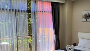 Quarto de hotel com cama, uma mesinha e vista para o exterior através de cortinas leves.