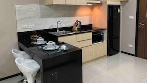Cozinha moderna com bancada preta, forno embutido e pia. Há uma pequena ilha com uma cadeira branca e um conjunto de chá. A cozinha possui armários bege e fogão preto.