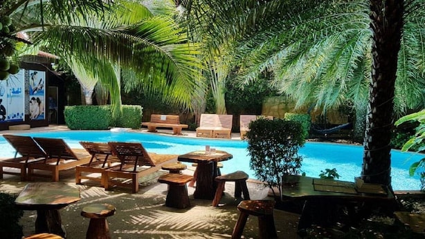 Uma área de piscina com espreguiçadeiras, mesas de madeira e uma rede.