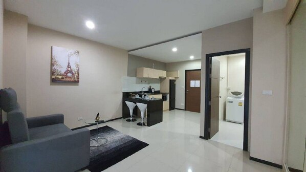 A modern living room with a sofa, a coffee table, and a painting of the Eiffel Tower. There is a kitchen area with cabinets and a refrigerator. - NBC  Nai Harn Beach Condo ISI (Rawai)