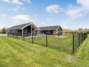A two-story building with a dark exterior, a covered patio area, and a fenced playground with a slide and swings. - Luxury Retreat by Beach - By Traum Ferienwohnungen (Idestrup)
