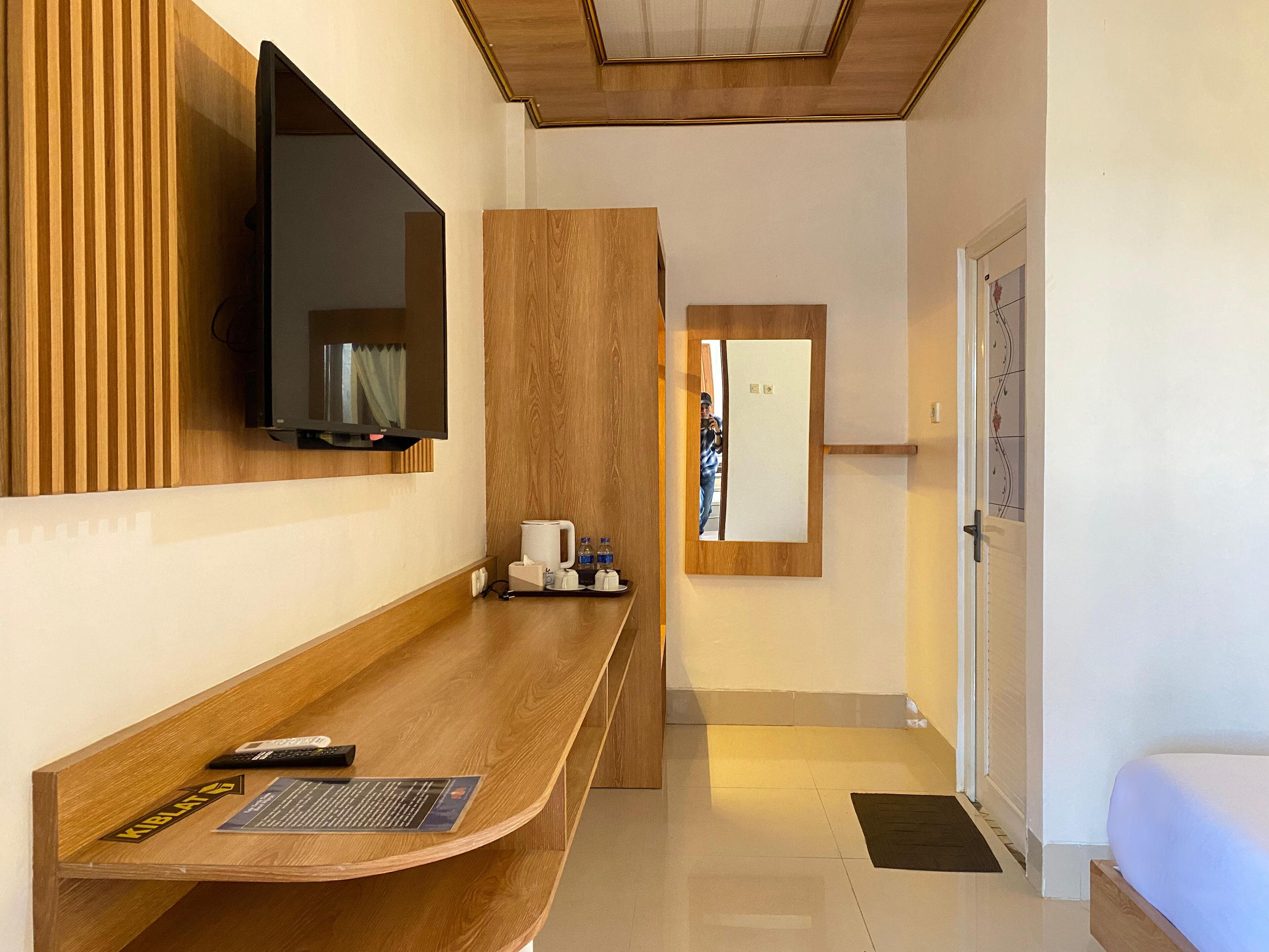A modern hotel room with a wooden reception desk, a mounted TV, a small seating area, and a door with a mirror.
