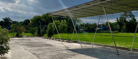 A covered outdoor area with a concrete floor, surrounded by greenery and a field in the background.