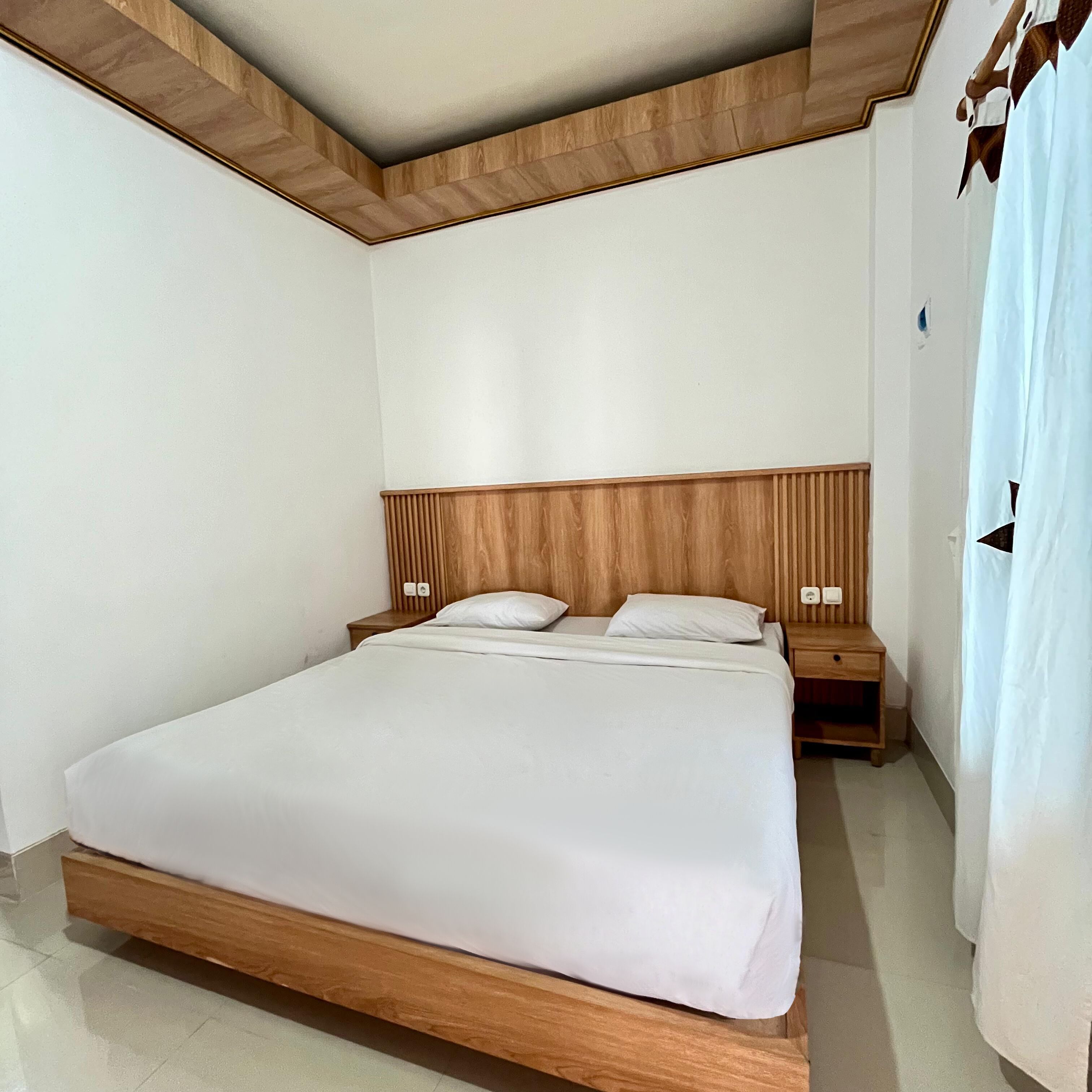 A neatly made bed with a wooden headboard, two bedside tables, and a white curtain.
