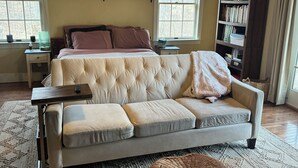 Une chambre à coucher avec un lit doté d’une tête de lit capitonnée, un canapé, un petit pouf, une bibliothèque et un ventilateur de plafond.