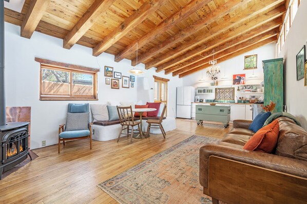 A cozy living room with a fireplace, wooden ceiling beams, a dining area, and a kitchen. - Stunning Artful Adobe - Super Walkable Location (Santa Fe)