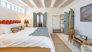 Un dormitori amb un llit gran, un panell de fusta al capçal, un mirall fixat a la paret, una taula petita amb una planta i un quadre a la paret.