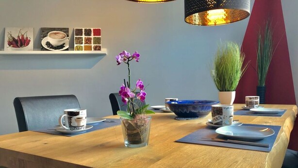 A wooden dining table with a coffee cup, a glass vase with purple flowers, a blue bowl, and a potted plant. - Holiday Apartment Erlebnisrhoen (Ehrenberg)