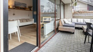 Ein zeitgemäßer Wohnbereich mit Glastür und Zugang zu einer Außenpatio. Innen befindet sich eine weiße Couch, eine Holzbank und ein Esstisch mit Stühlen. Die Patio hat einen Ziegelboden und Blick ins Freie.