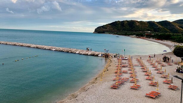 Una playa con filas de tumbonas y sombrillas, un rompeolas rocoso y un paisaje montañoso al fondo.
